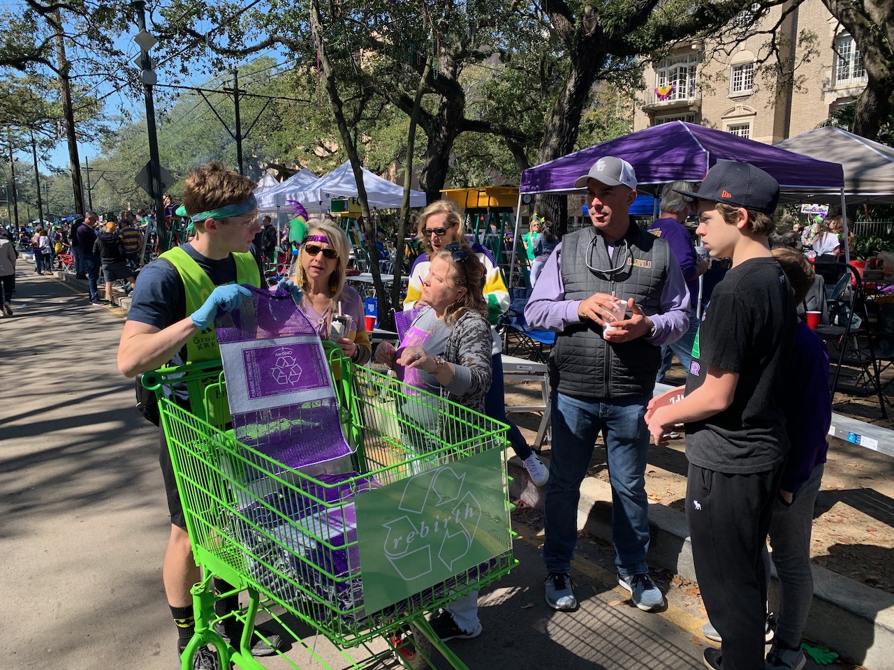 Year-one volunteer pushing a green shopping cart with a 'rebirth' recycling sign down St. Charles, neutral ground tents in the background