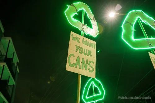 Glowing neon green recycle-symbol signs over a hand-painted cardboard sign that reads WE WANT YOUR CANS, at night during the Recyclists Marching Krewe parade