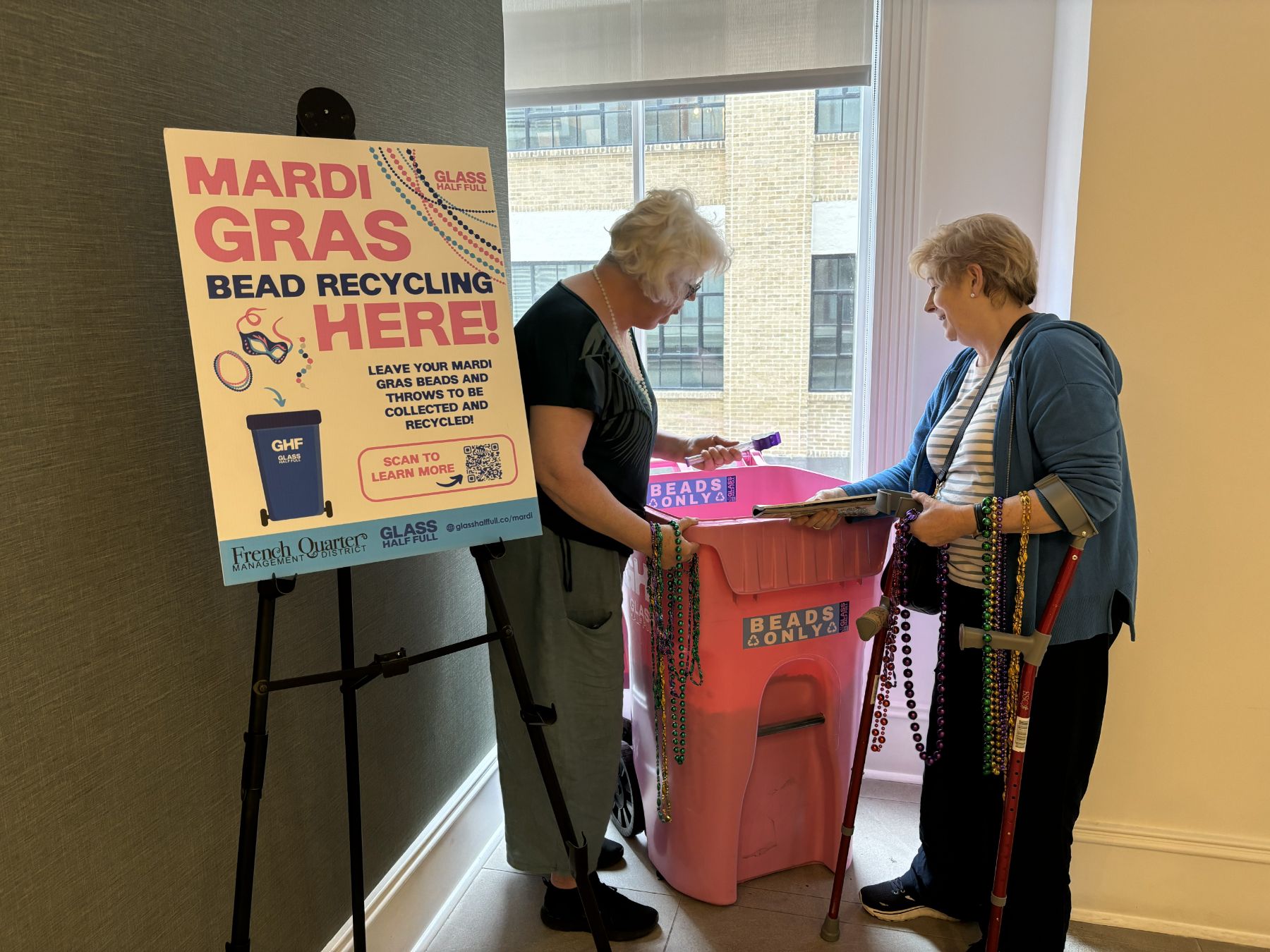Two hotel guests dropping Mardi Gras beads into a pink Glass Half Full bin next to a MARDI GRAS BEAD RECYCLING HERE easel sign