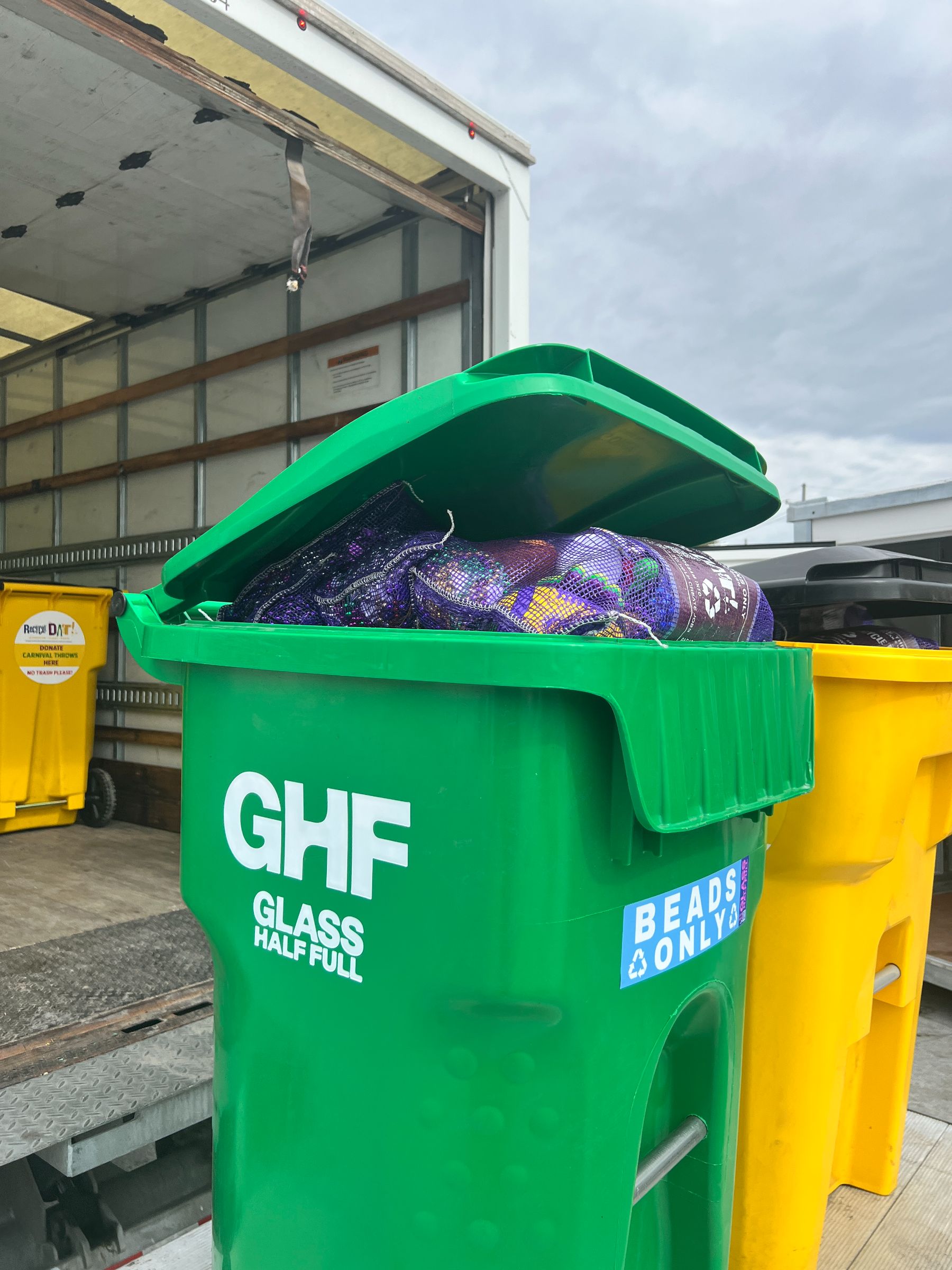A green Glass Half Full bin filled with bagged beads being loaded into a box truck next to a yellow RecycleDAT bin