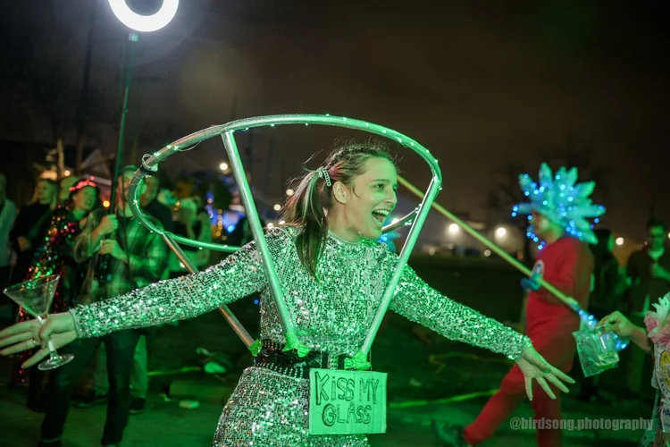Krewe member in a sequined dress with an oversized martini-glass headpiece, sign reading KISS MY GLASS